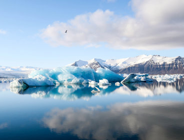 A white a blue glacier, seen emerging from a very still ocean. The ocean is so still that it is reflecting the blue sky and clouds above. Mountains are in the background