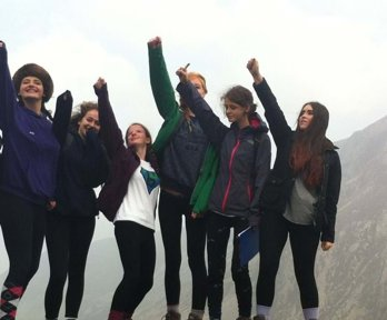 A group of young women standing on a rock in the outdoors with their arms reaching int the air, clearly enjoying geography