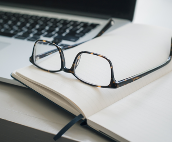 A pair of glasses, open laptop, empty notebook and a pen messily lay on top of each other on a plain grey surface.