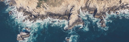 An aerial image of a rocky coastline edge, with mud and vegetation on the cliffs and waves breaking around the cliff foot.