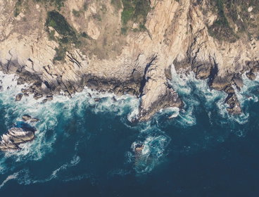 An aerial image of a rocky coastline edge, with mud and vegetation on the cliffs and waves breaking around the cliff foot.