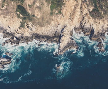 An aerial image of a rocky coastline edge, with mud and vegetation on the cliffs and waves breaking around the cliff foot.