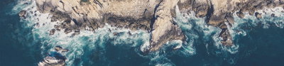 An aerial image of a rocky coastline edge, with mud and vegetation on the cliffs and waves breaking around the cliff foot.