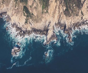 An aerial image of a rocky coastline edge, with mud and vegetation on the cliffs and waves breaking around the cliff foot