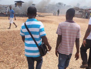 Three people walking in a row on wood chips towards a mound of waste. Smoke is rising from the mound where waste is on fire.