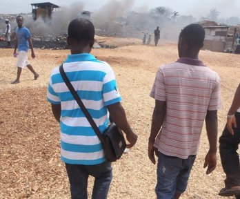 Three people walking in a row on wood chips towards a mound of waste. Smoke is rising from the mound where waste is on fire.