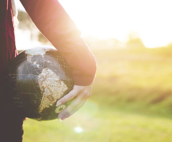 A person holding a small globe in their hand, down by their side. The sun is low in the sky and the light is bouncing off the top of the globe