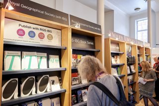 Person with shoulder length grey hair and backpack standing in front of a bookcase browsing journals and books from RGS-IBG publications.