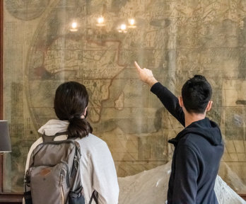 The people stand in from of a large wall mounted map with their back to the camera as they look and point at the map