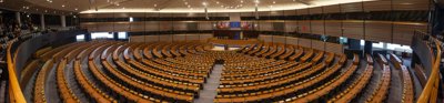 An elevated view of a very large auditorium filled with brown wood desk and black office chairs. The European Union flag is pinned up behind the podium inbetween the flags of all the EU nation drapped on flag polls.