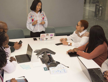 Six people sit around a white table in a board room listening to a seventh person at standing at the head of the table talk.