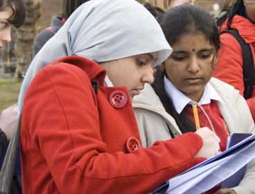 Female students working outsdie, one is holding a clipboard and making notes while the other is watching what she is writing