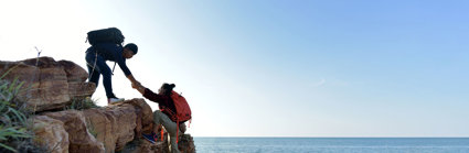 Two hikers on a seaside cliff on a sunny day. One is assisting the other by taking their hand and pulling them up. 