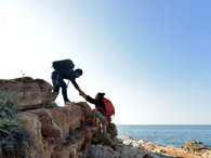 Two hikers on a seaside cliff on a sunny day. One is assisting the other by taking their hand and pulling them up. 