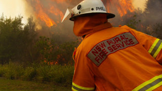 Two firemen are fighting a fire by the side of the road. Their uniforms are orange and their hose is yellow. The flames are extremally high and there is smoke