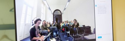 A digital screen showing a room with seated people