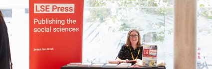 Photograph of the LSE Press stall at the RGS-IBG Conference with a white lady sitting behind the desk. A red banner reads 'LSE Press: Publishing the social sciences'.