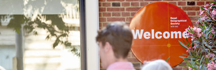 People walk into the RGS at Exhibition Road. An orange welcome sign is focused in on.