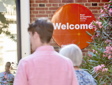 People walk into the RGS at Exhibition Road. An orange welcome sign is focused in on.