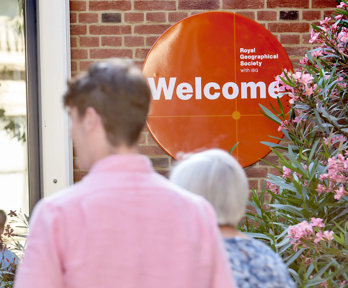 People walk into the RGS at Exhibition Road. An orange welcome sign is focused in on.