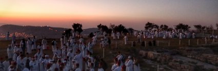 A large group of people gathered for a ceremony on a mountaintop as the sun is setting. 