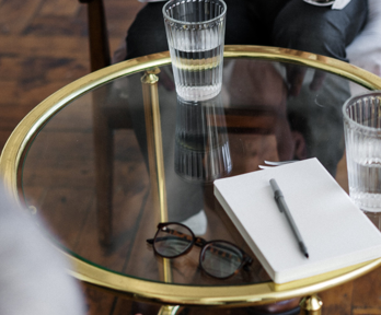 A notepad, pen and glasses sit on a glass table alongside two glasses of water. The two smartly dressed individuals sit buy the table yet slightly out of view