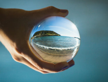 A hand cups around a glass ball. Inside the glass ball is an image of a beach with waves splashing and the sun shining down on the water. 