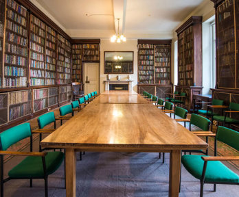 A large wooden table setup for a boardroom meeting in the Lowther Room at the Society. The table is surrounded by large bookcases full of books.