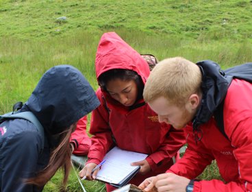 Three young people stand in a shallow stream completing fieldwork research while wearing outdoor clothing and holding clipboards.