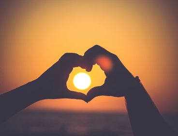 Silhouette of a persons hands making a heart shape around the sun