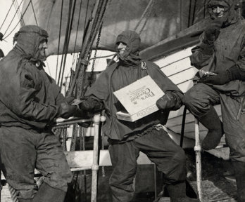 Black and white photograph of three people on a boat dressed in waterproof gear. They're smiling and eating chocolate from a box of Fry's Chocolate.