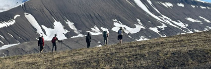 Five people walking in a line through a mountainous landscape. The mountain slope is largely bare with patches of snow remaining. 