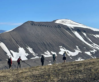 Five people walking in a line through a mountainous landscape. The mountain slope is largely bare with patches of snow remaining. 