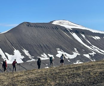 Five people walking in a line through a mountainous landscape. The mountain slope is largely bare with patches of snow remaining. 