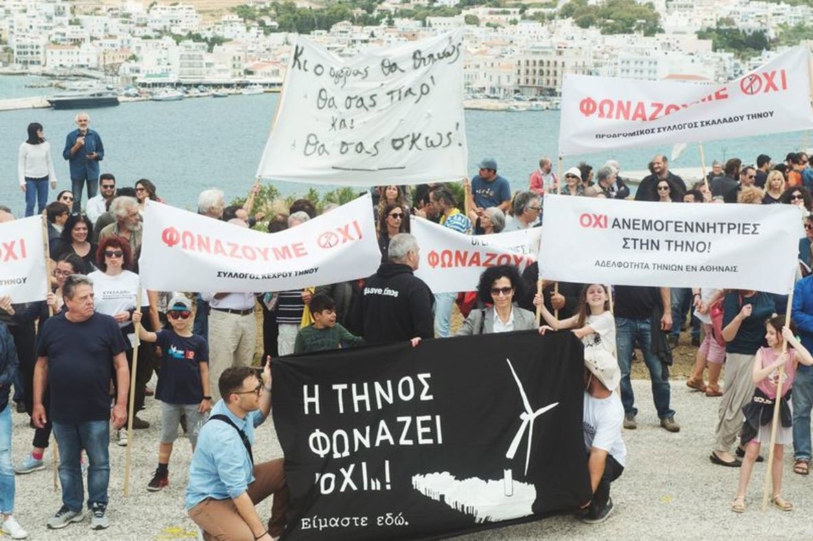 A large group of people holding up banners in front of a marina. The front banner has an image of a broken wind turbine.