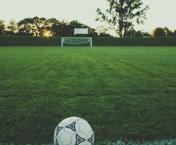 A football set on a green pitch with a goal in the background. It is dusk and the sun i setting behind the trees, which are behind the goal