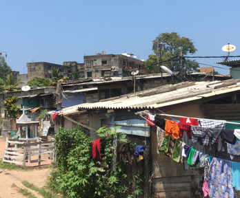 A person walks on a unpaved dirt road encroached on by grass and other vegetation. To the right makeshift buildings with aluminium roofs fill out the road side. clothing hangs on a washing line between two.