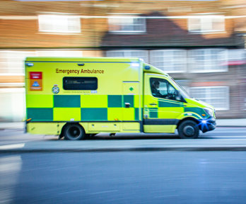 An ambulance driving at speed. The background of the picture, which is a residential street, is blurry to indicate the speed at which the vehicle is travelling.