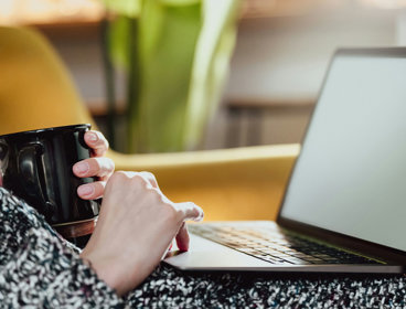 Person sat in a chair, holding a mug with a laptop on their lap. 