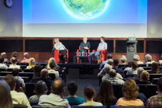 Three men sit in red chairs on the stage of a lecture theatre having a discussion with the image of a globe above them on a screen. The back of the heads of the audience can be seen in the foreground.