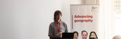 A speaker is presenting behind a lecturn. A poster next to him reads 'advancing geography'.