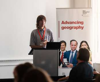 A speaker is presenting behind a lecturn. A poster next to him reads 'advancing geography'.