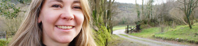 Headshot of Belinda Kirk. In the background is a forest and unpaved swirling countryside road. 