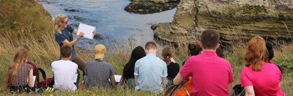 A teacher is standing at the top of a cliff, overlooking a rocky beach cove, holding a clipboard and showing a group of students something