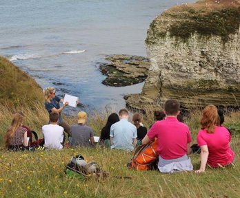 A teacher is standing at the top of a cliff, overlooking a rocky beach cove, holding a clipboard and showing a group of students something
