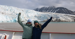 Two people on a boat in front of a glacier with their arms in the air. 