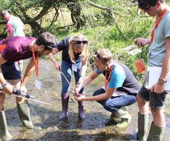 A group of teachers are standing in a river, all wearing wellies, measuring the depth of the river using a stick.