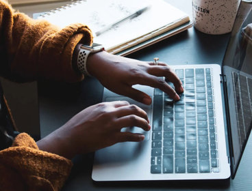 A person at a desk typing on a laptop with a notebook and mug to one side.