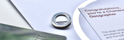 A silver circle lapel pin, a symbol of Chartered Geographer status, next to a card that says 'Congratulations, you're a Chartered Geographer'. 