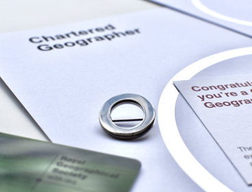 A silver circle lapel pin, a symbol of Chartered Geographer status, next to a card that says 'Congratulations, you're a Chartered Geographer'. 